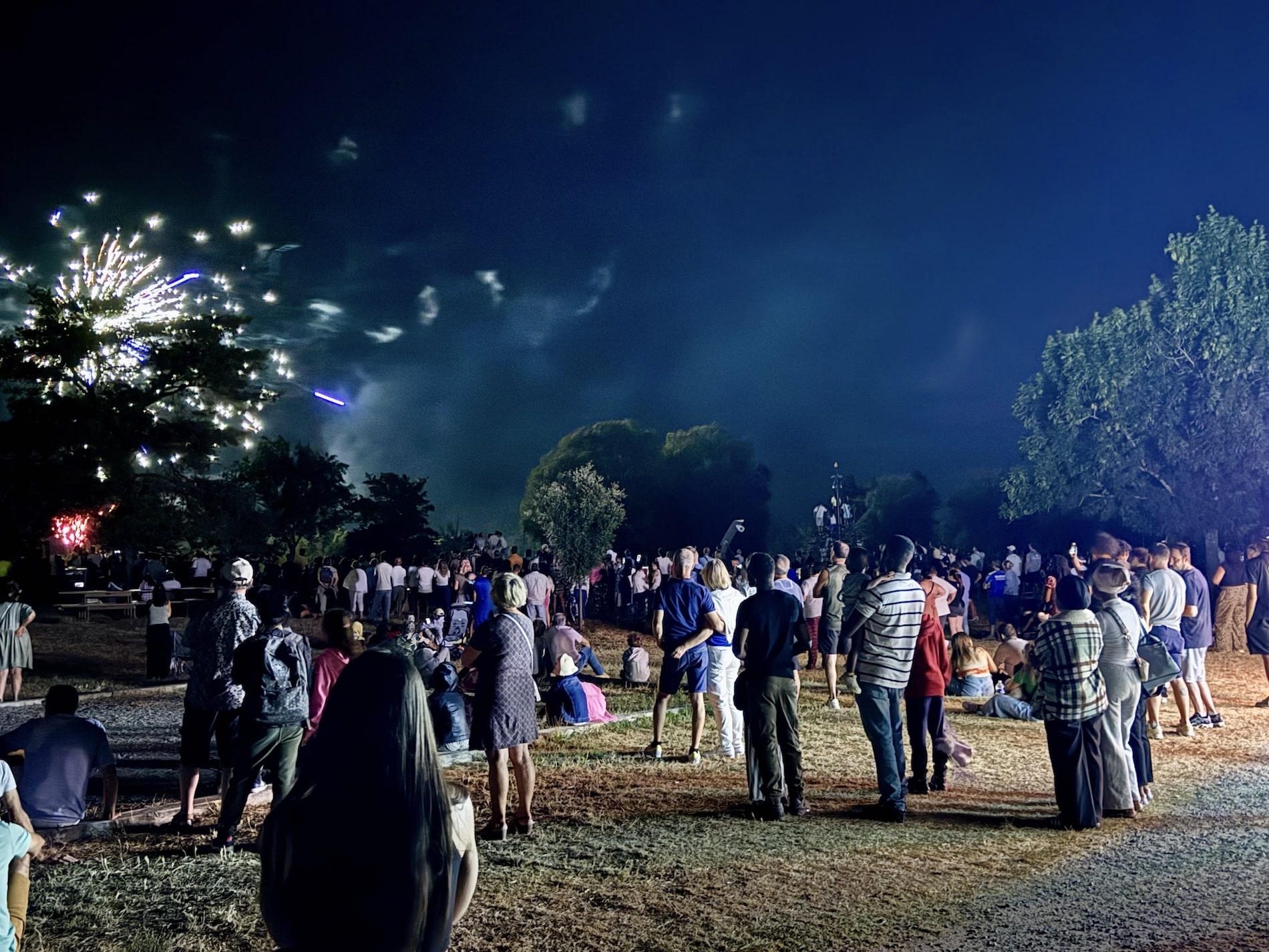 Une foule regarde un feux d'artifice durant une nuit d'été. Une explosion blanche se devine à la gauche de l'image.